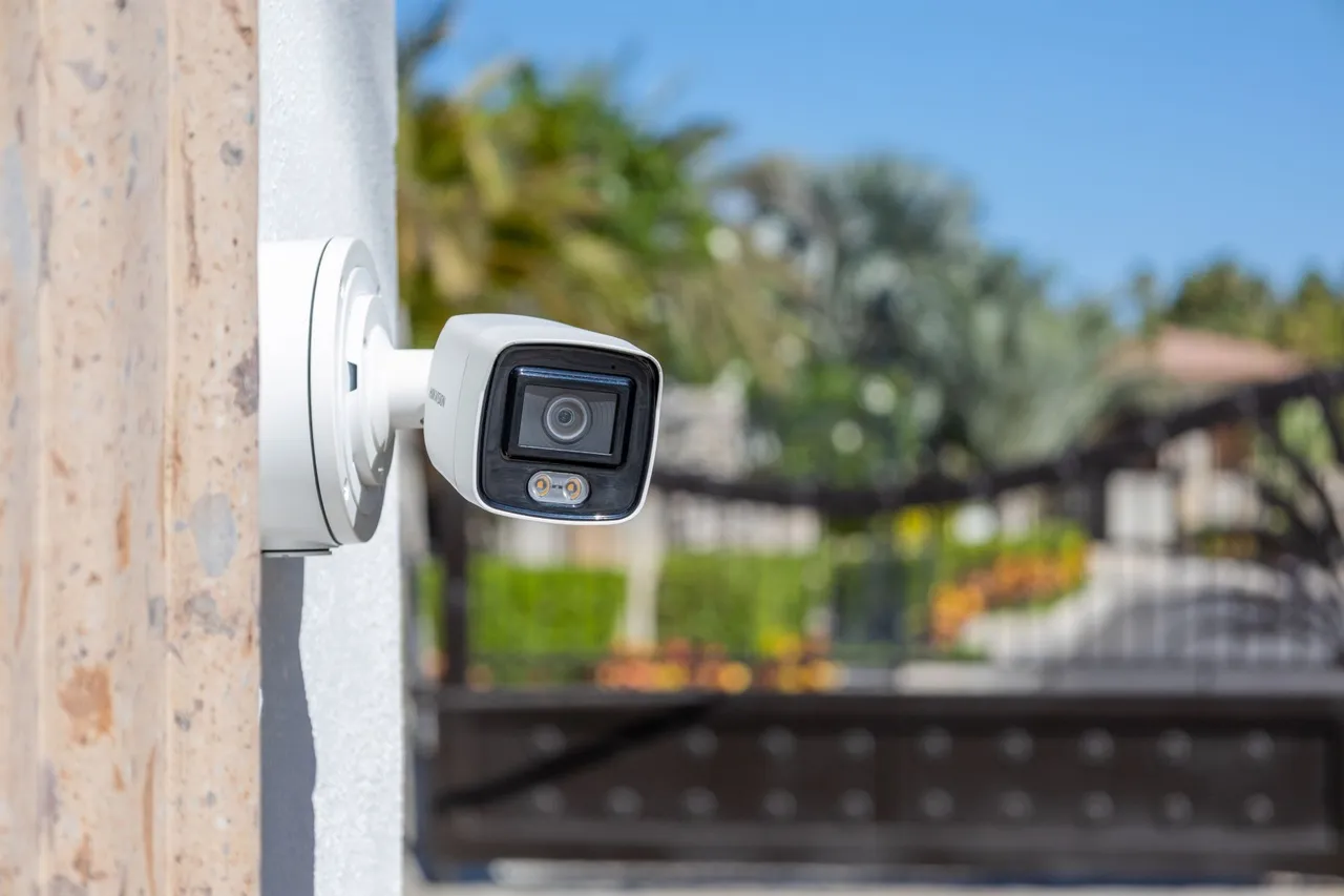 Bullet camera on a column, with a gate and palm trees out of focus behind